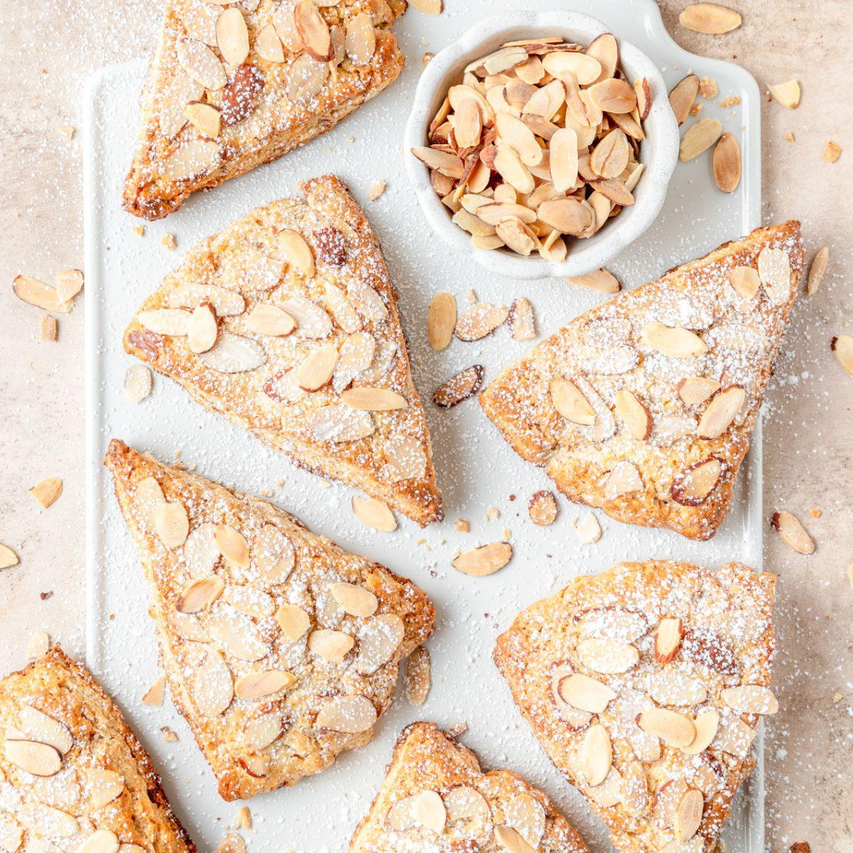 Triangle shaped baked scones, topped with sliced almonds. A bowl filled with sliced almonds.